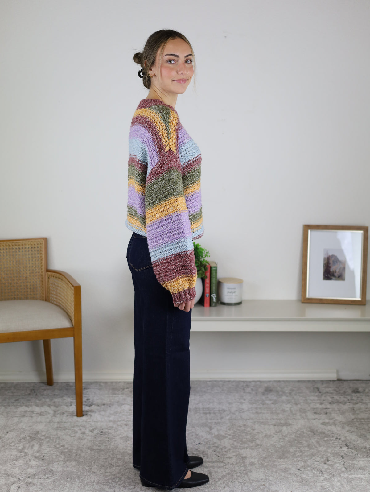 A person wears a cozy multi-color Reign Striped Knit Sweater in shades of purple, green, yellow, and red, paired with dark blue wide-leg jeans.