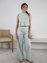 Wearing the Myra Gingham Print Ruffled Set—a sleeveless, sage gingham two-piece with a ruffled peplum top and matching high-waisted wide-leg pants—a woman stands indoors beside a wooden chair on a light carpet, set against a neutral background.