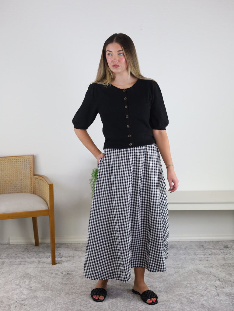 A woman stands indoors wearing the Monica Short Sleeve Cardigan in black, styled with a black and white gingham skirt and black slide sandals. In the background, there is a wooden chair and a white bench set against a plain white wall.