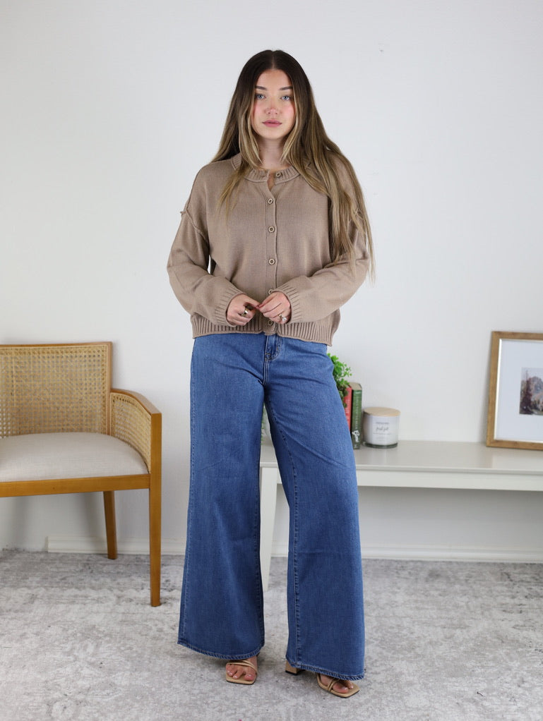 A woman wearing a tan, raw edge Jude Raw Edge Cardigan with a button-front and wide-leg blue jeans.
