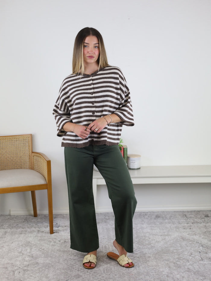 A person stands indoors wearing a brown and white striped top, dark green wide-leg pants, and tan sandals. With straight, shoulder-length hair, they pose in a minimalist room featuring a chair and a shelf in the background. Their effortless look is completed with the Lottie Striped Roll Neck Cardigan—an ideal transitional layering piece for changing seasons.