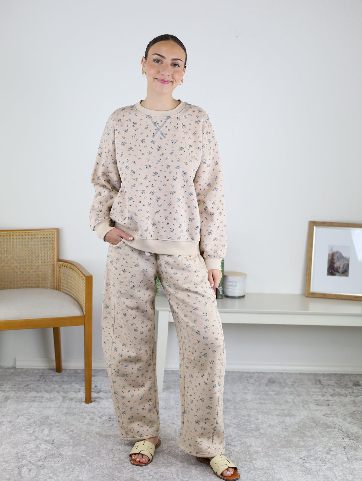 A woman stands indoors wearing the Journey Floral Boxy Crewneck Lounge Set—a matching beige khaki two-piece with a floral sweatshirt and pants. She pairs the outfit with sandals and earrings, smiling with one hand in her pocket. A chair and a white console table can be seen in the background.
