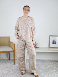 A woman stands indoors wearing the Journey Floral Boxy Crewneck Lounge Set—a matching beige khaki two-piece with a floral sweatshirt and pants. She pairs the outfit with sandals and earrings, smiling with one hand in her pocket. A chair and a white console table can be seen in the background.