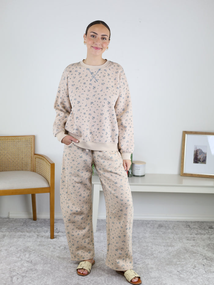 A woman stands indoors wearing the Journey Floral Boxy Crewneck Lounge Set—a matching beige khaki two-piece with a floral sweatshirt and pants. She pairs the outfit with sandals and earrings, smiling with one hand in her pocket. A chair and a white console table can be seen in the background.