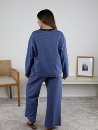 A woman stands indoors facing away, wearing the Misty Striped Relaxed Top & Wide Leg Pants. A chair is to her left, and a framed photo rests on a white shelf against a plain wall.