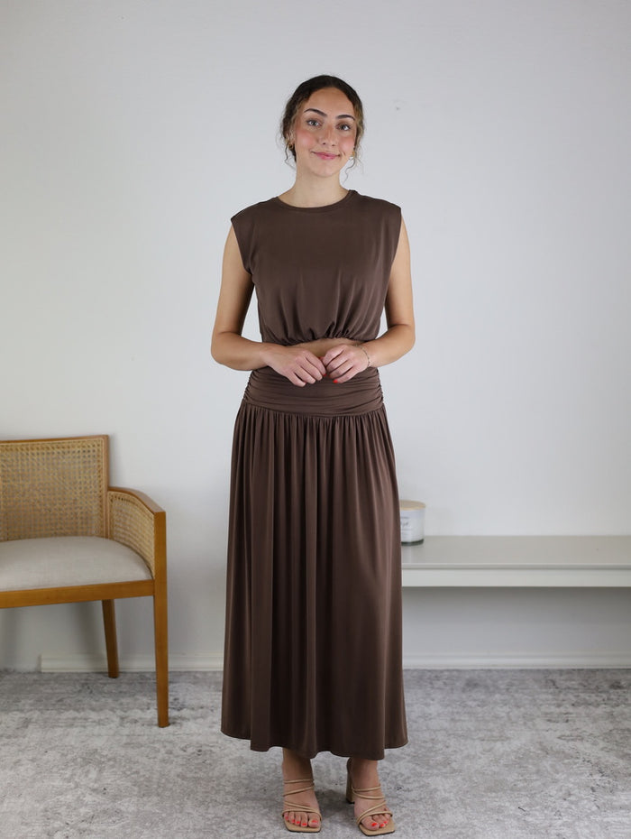 A woman stands indoors wearing the Harlow Bubble Top w/ Skirt, styled with tan sandals and a subtle smile—an effortlessly chic choice for a night out. A wooden chair and a low white bench rest against the plain white wall in the background.