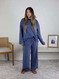 A woman stands indoors wearing the Misty Striped Relaxed Top & Wide Leg Pants set. A wooden chair and a framed picture are displayed on the shelf beside her.