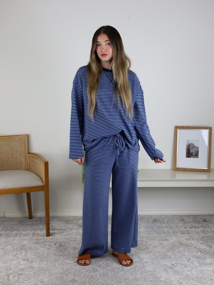 A woman stands indoors wearing the Misty Striped Relaxed Top & Wide Leg Pants set. A wooden chair and a framed picture are displayed on the shelf beside her.