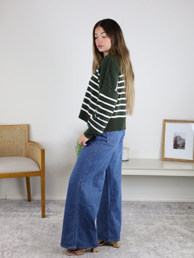 A person wearing an oversized forest green and white striped Noa Striped Sweater with wide-leg blue jeans.