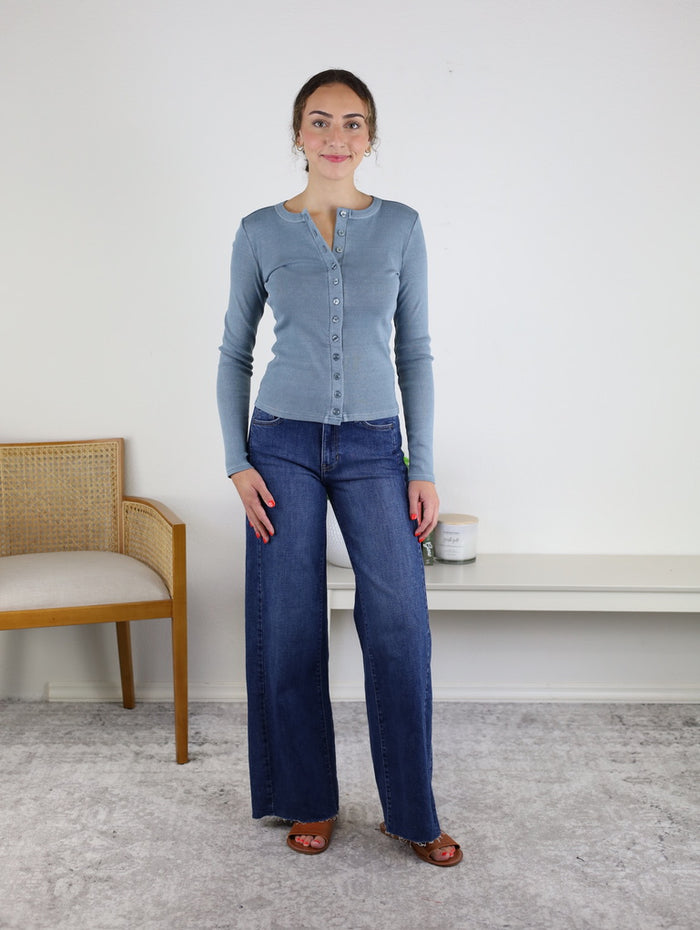 A woman stands indoors on a light rug, wearing wide leg denim jeans—the Maisey Raw Hem Side Panel Wide Leg—and a light blue button-up top. She has brown sandals on and is smiling. A wooden chair and two candles sit in the background.