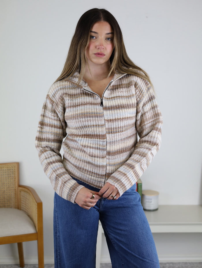 A person wears a Romy Hooded Cardigan Sweater, a cozy knit hoodie with a zip front and a ribbed texture, featuring horizontal stripes in shades of beige, brown, and white.