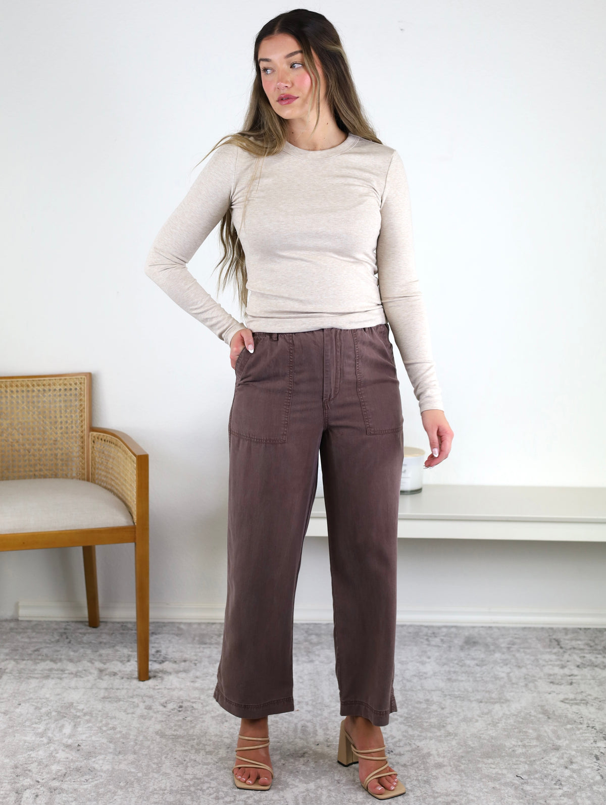 A woman stands indoors, gazing to the side with one hand in her pocket, wearing a beige long-sleeve top paired with the Marbella Wide Leg Tencel Pant in a rich brown shade and heeled sandals. A wooden chair and bench are visible in the background.