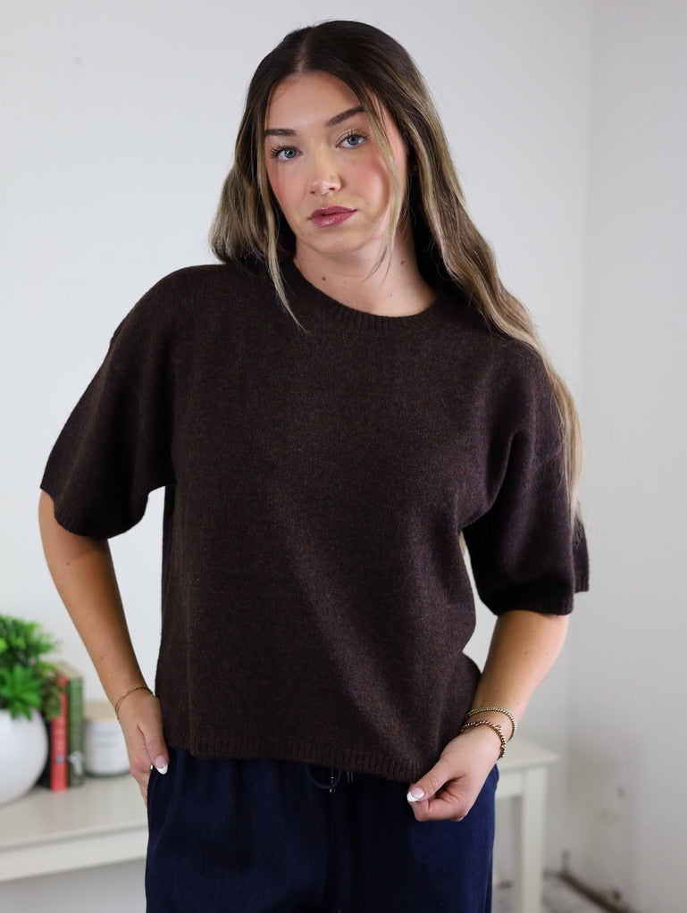 A woman with long brown hair models the West Raw Edge Sweater—a relaxed, short-sleeve pullover in a dark brown cotton-acrylic blend—while posing indoors in front of a white wall and a small table topped with a potted plant and books.