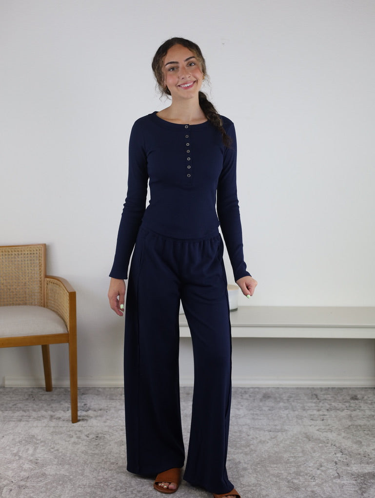 A smiling woman with long brown hair in a braid wears the Statler Lounge Set—featuring a navy henley top and wide-leg pant—while standing indoors on a gray rug near a wooden chair and white bench.