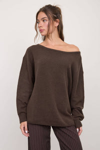 A woman wearing The Workshop Studio’s Boat Neck Oversized Knit Pullover Sweater in Red Wood and brown pinstriped pants stands in front of a plain light background, looking to her left.