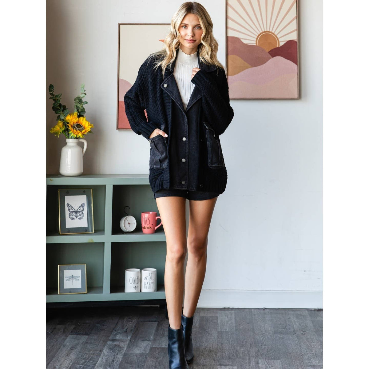 A woman stands indoors wearing the Veveret - Knit Body Denim Layerd Jacket: Cream / Denim / L over a white turtleneck, black skirt, and ankle boots, posing with one hand in her pocket in front of bookshelves and flowers.