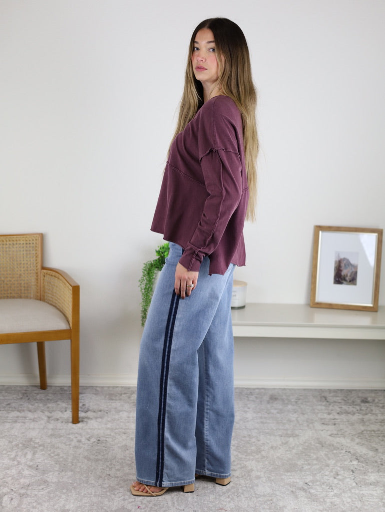 Woman standing indoors, wearing an Ester Raw Seam Henley in maroon and light blue wide-leg jeans with a dark stripe down the side. She faces sideways on a light grey carpet; chair and plant behind her.