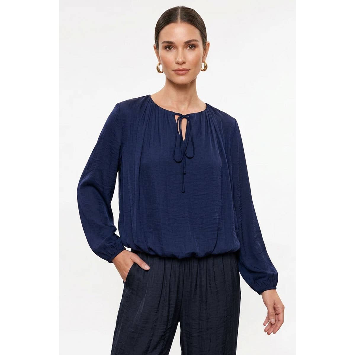 A woman wearing a Naked Zebra - Long Sleeve Draped Bubble Hem Blouse in wine, featuring a long-sleeved satin design with a tie neckline and a gathered, draped bubble hem.