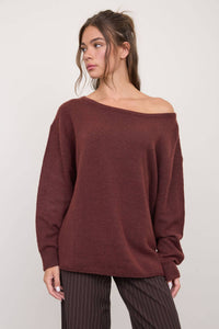 A woman stands against a plain light background, looking to the side, wearing The Workshop Studio’s Boat Neck Oversized Knit Pullover Sweater in Red Wood with a loose, off-the-shoulder fit, paired with brown pinstripe pants.