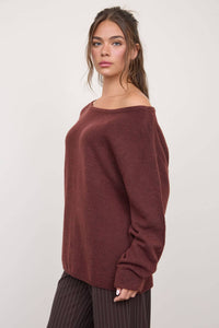 A woman with long brown hair stands sideways against a plain light background, looking at the camera with a neutral expression. She wears The Workshop Studio - BOAT NECK OVERSIZED KNIT PULLOVER SWEATER in Red Wood, styled loosely off-the-shoulder with an oversized fit, paired with dark pinstriped pants.
