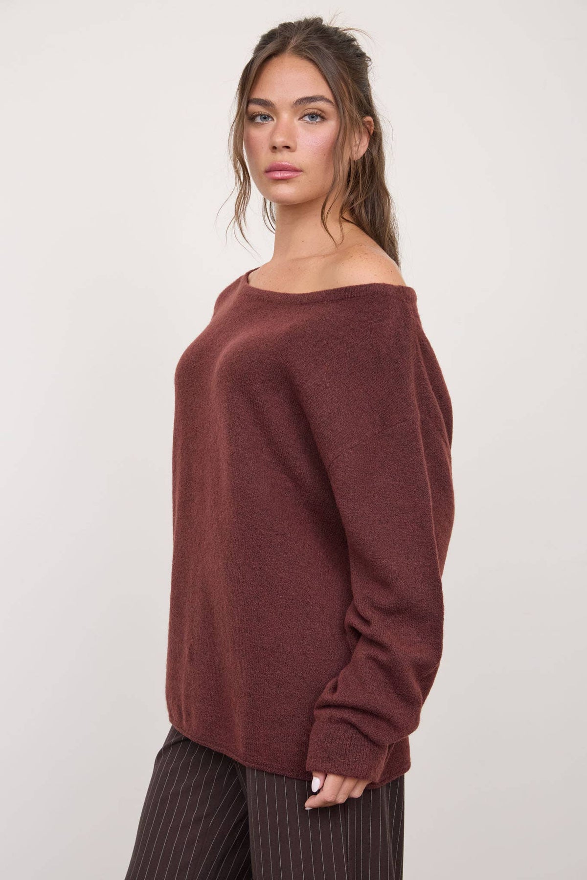 A woman stands against a plain light background, looking confidently at the camera with relaxed hair, wearing The Workshop Studio - BOAT NECK OVERSIZED KNIT PULLOVER SWEATER in Oatmeal paired with brown pinstripe pants.
