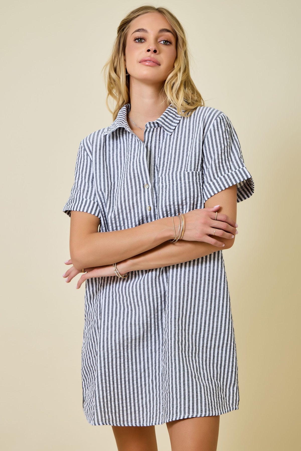 A woman stands against a plain background, arms loosely crossed and wearing a slight smile, dressed in the Renee Striped Seersucker Shirt Dress—a short-sleeve, button-up design featuring blue and white stripes and crafted from a soft poly-cotton blend.
