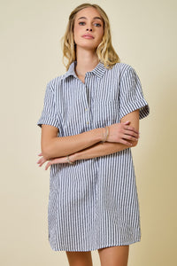 A woman stands against a plain background, arms loosely crossed and wearing a slight smile, dressed in the Renee Striped Seersucker Shirt Dress—a short-sleeve, button-up design featuring blue and white stripes and crafted from a soft poly-cotton blend.