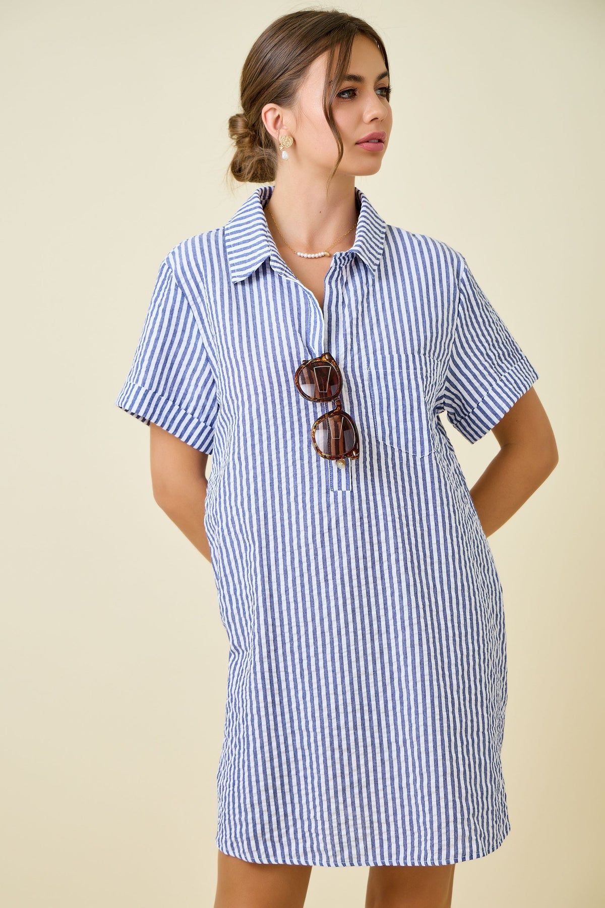 A woman wears the Renee Striped Seersucker Shirt Dress—a blue and white vertical striped style perfect for summer—while holding brown sunglasses and looking to the side. Her hair is styled in a low bun, and she completes her look with earrings and a necklace.