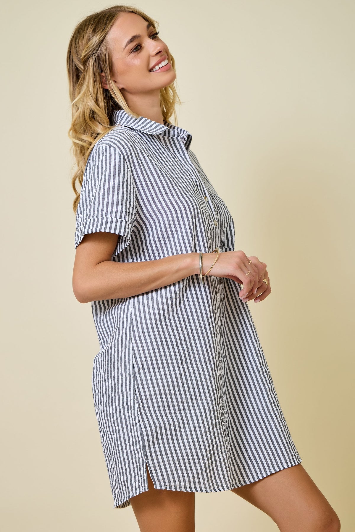 A smiling woman stands sideways against a beige background wearing the Renee Striped Seersucker Shirt Dress—a short-sleeve, black and white striped dress crafted from a poly-cotton blend.