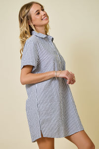 A smiling woman stands sideways against a beige background wearing the Renee Striped Seersucker Shirt Dress—a short-sleeve, black and white striped dress crafted from a poly-cotton blend.
