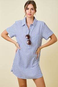 A woman stands with hands on hips, holding sunglasses, wearing the Renee Striped Seersucker Shirt Dress—a blue and white vertically striped summer dress made from a soft poly-cotton blend. The background is light beige.