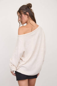 A woman with long brown hair styled in a loose bun wears The Workshop Studio Boat Neck Oversized Knit Pullover Sweater in oatmeal off one shoulder, paired with a black skirt. She stands against a plain light background, gazing down over her shoulder.