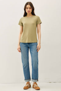 A woman stands against a plain background, wearing the Fern Basic Bamboo Tee in light olive green with a relaxed fit, paired with blue cuffed jeans and brown sneakers. She has long brown hair and a neutral expression.