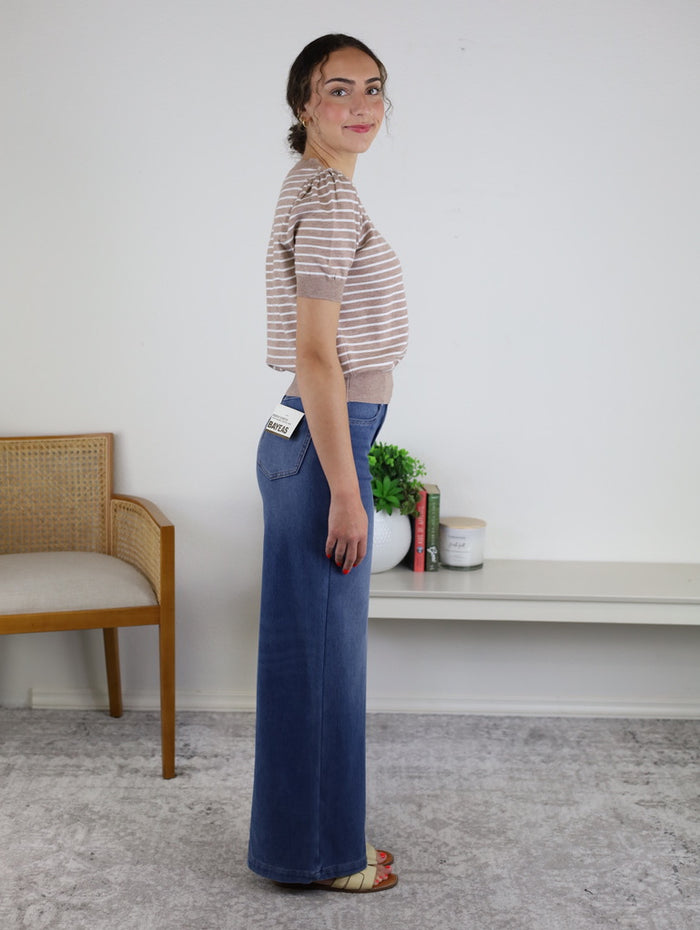 A woman stands sideways, wearing the Ace Puff Striped Sweater—a lightweight knit with short sleeves and stripes—paired with wide-leg blue jeans featuring a visible tag and sandals. She is in a simple, modern room with a chair, a white shelf, books, and a plant.