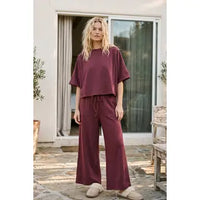 A woman with long blonde hair and a relaxed expression stands outside in front of glass doors, wearing the Heather Brushed Stretch Boxy Top & Wide Leg Pant Set—an effortlessly cozy maroon loungewear ensemble featuring a loose-fitting boxy top and wide leg pants. She completes her look with cream-colored slippers.