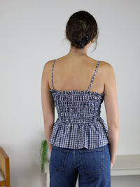 A woman with dark hair in a bun, dressed in the Lonnie Smocked Gingham Peplum Top and blue jeans, stands with her back to the camera in a minimal room featuring a chair and a plant.
