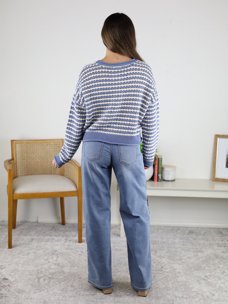 Woman stands indoors with her back to the camera, wearing the Bellamy Textured Striped Crochet Cardigan and light blue wide-leg jeans. She is in a room with a beige chair, white shelf, books, and framed art.