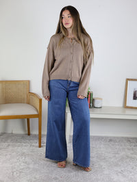 A woman wearing a tan, lightweight Jude Raw Edge Cardigan with raw-edge detailing and wide-leg blue jeans.