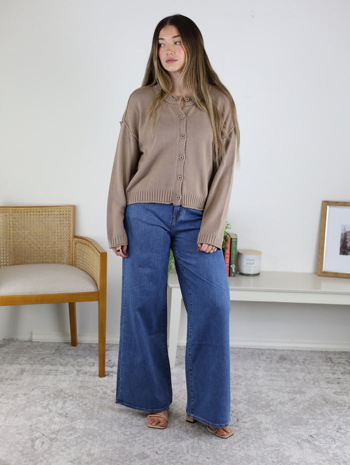 A woman wearing a tan, lightweight Jude Raw Edge Cardigan with raw-edge detailing and wide-leg blue jeans.