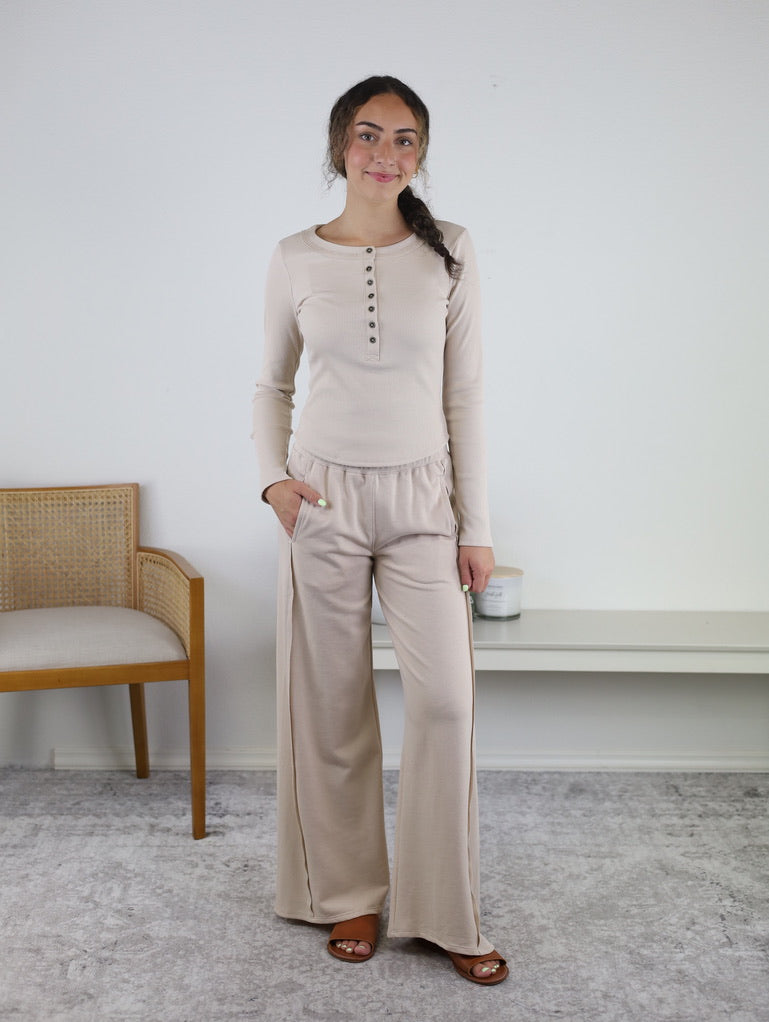 A woman stands indoors wearing the Statler Lounge Set—a beige ensemble featuring a henley top with buttons and matching wide-leg pants. She strikes a relaxed pose with one hand in her pocket, while a chair and a white shelf appear in the background.