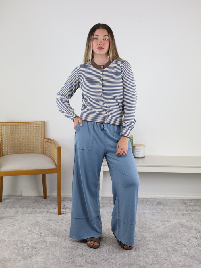 A woman stands indoors wearing the Hazel Striped Cardigan, a cozy and lightweight polyester knit layer, paired with loose blue pants. She has one hand in her pocket and is standing on a light gray rug, with a chair and a white bench behind her.