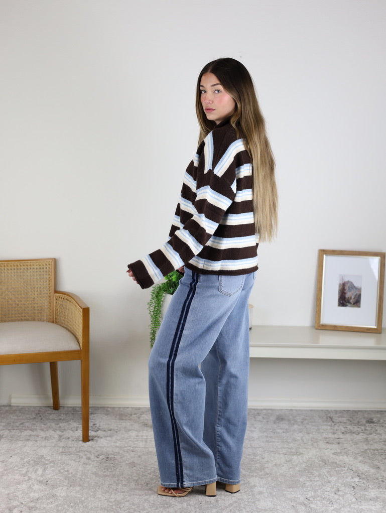 A person wearing a brown, white, and blue Remy Multicolor Rolled Neck Knit Cardigan with light-wash wide-leg jeans featuring a dark side stripe.