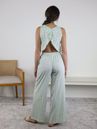 A woman stands facing away, wearing the Myra Gingham Print Ruffled Set—a light green and white checkered sleeveless peplum top with a crisscross back and matching wide-leg pants. She is near a tan chair and white bench in a simple room.
