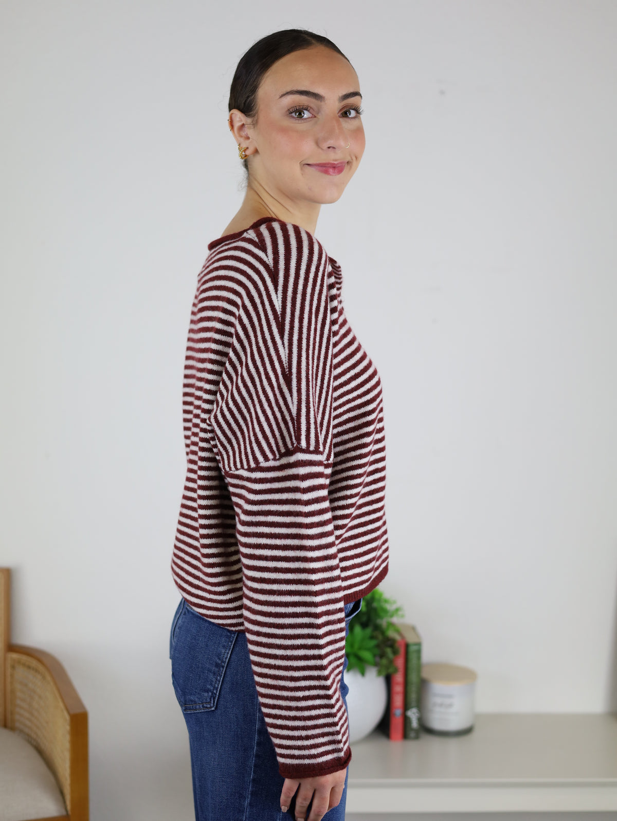 A woman stands sideways indoors, smiling slightly as she wears the Maren Striped Cardigan in a relaxed fit with blue jeans. In the background, there is a chair, small plant, books, and a candle on a table.