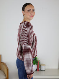 A woman stands sideways indoors, smiling slightly as she wears the Maren Striped Cardigan in a relaxed fit with blue jeans. In the background, there is a chair, small plant, books, and a candle on a table.