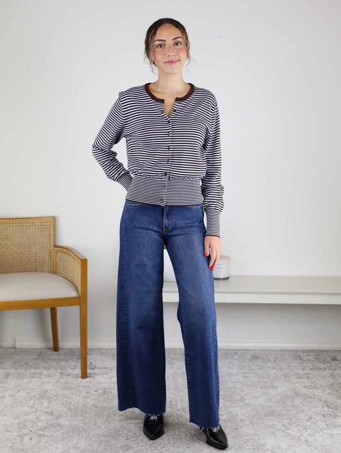 A woman stands indoors, smiling softly with one hand on her hip. She is wearing the Quinn Striped Cardigan paired with blue wide-leg jeans and black shoes. In the background, a wooden chair and bench are visible.