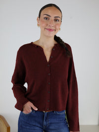 A woman with a long braid, dressed in the Beth Lightweight Cardigan and blue jeans, stands with one hand in her pocket against a plain white background. She is smiling slightly and looking at the camera.