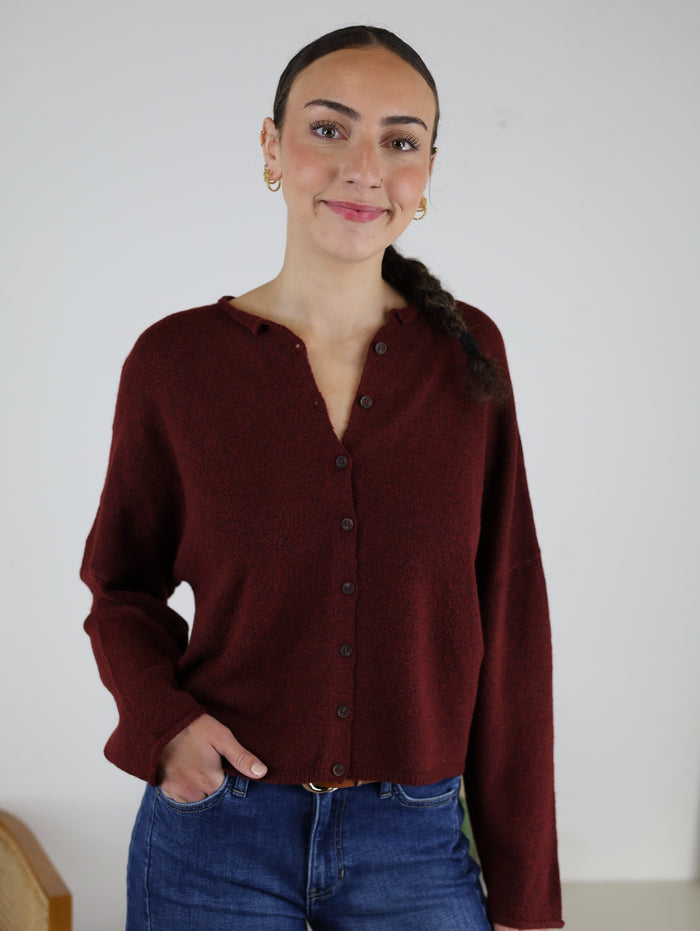 A woman with a long braid, dressed in the Beth Lightweight Cardigan and blue jeans, stands with one hand in her pocket against a plain white background. She is smiling slightly and looking at the camera.