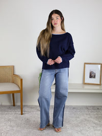A woman with long hair wears the Bell Contrast Boat Neck Sweater in navy, paired with light blue jeans, standing indoors on a light carpet next to a wooden chair and a shelf with decor.