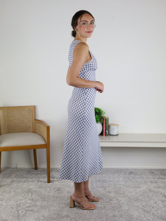 A woman stands sideways and smiles in a minimalist room with a wooden chair, white shelf, plant, and books. She wears the Tessa Sleeveless V-Neck Midi in blue and white checkered print with tan heeled sandals on a light-colored carpet.